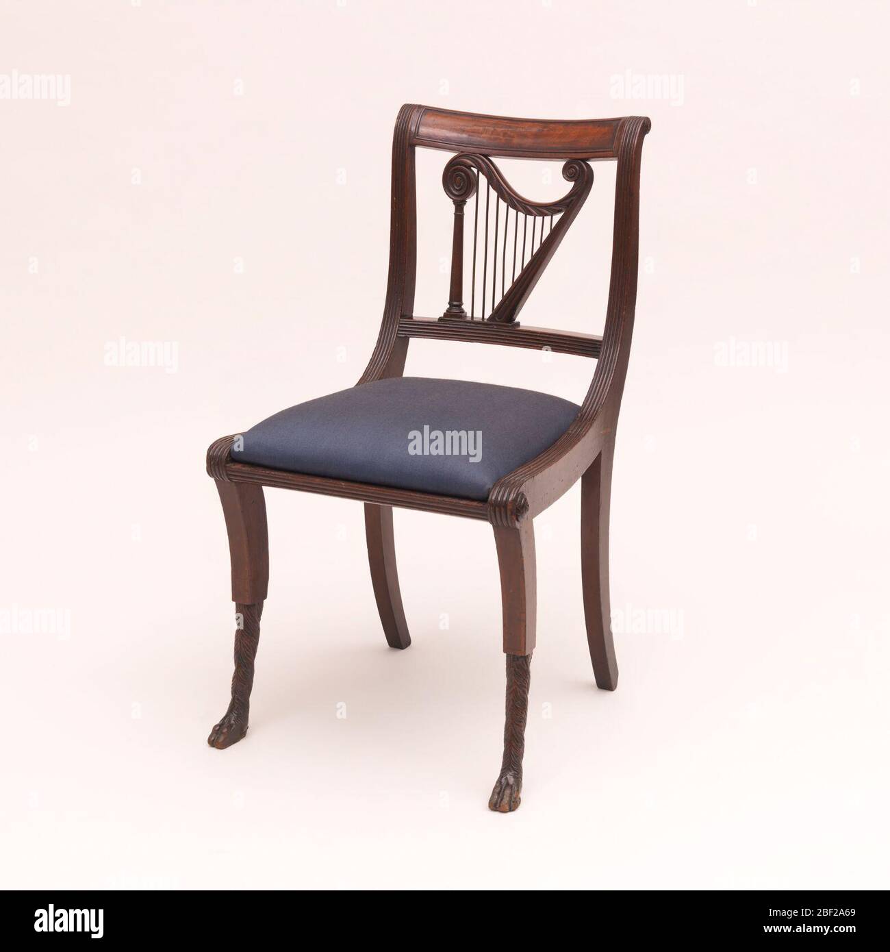 Side chair. The chair back with reeded posts; panelled top rail and reeded lower rail, enclosing the figure of a harp; out-curved front legs, lower half carved to depict fur-covered animals legs and paw feet; trapezoidal, dark blue slip seat in readed frame. Stock Photo