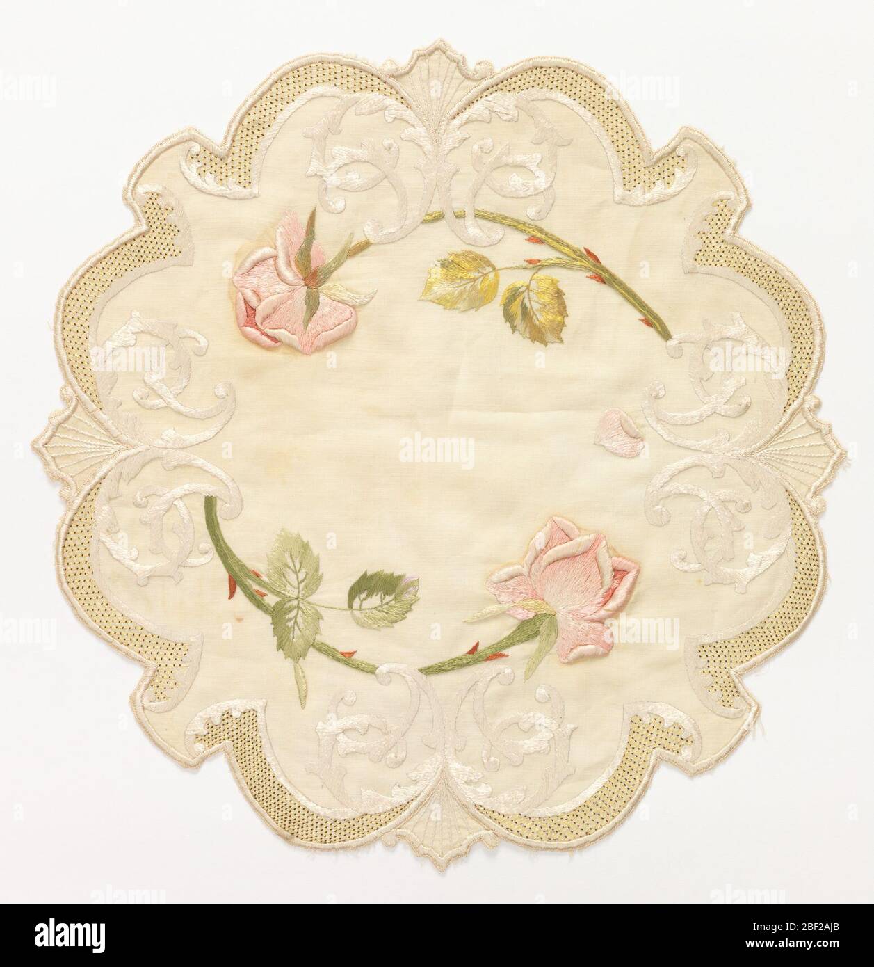 Table mat. Table mat of off-white linen, approximately round with scalloped edges. Embroidered in pale greens, pinks, red and off-white silks with a cipher entwined with two pink roses. Border of small yellow lattice with red dots. Stock Photo