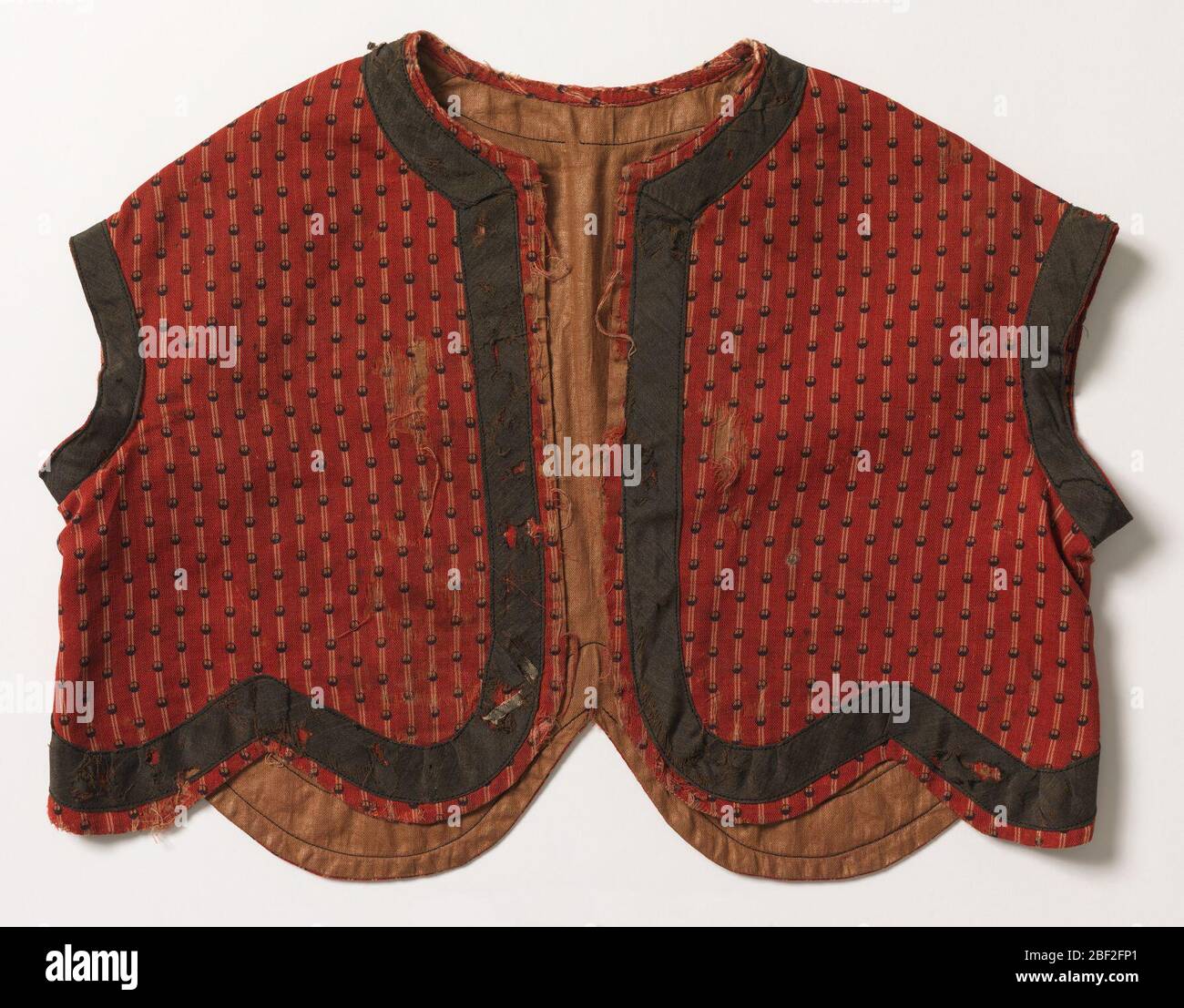 Vest. child's short jacket of red wool, striped in white and printed, over-stripe in small black dots. Roud neck, no sleeve, shaped, scalloped bottom. Trimmed with band of black wool around neck, arm hole and bottom, light brown glazed cotton lining. Machine sewn. Stock Photo