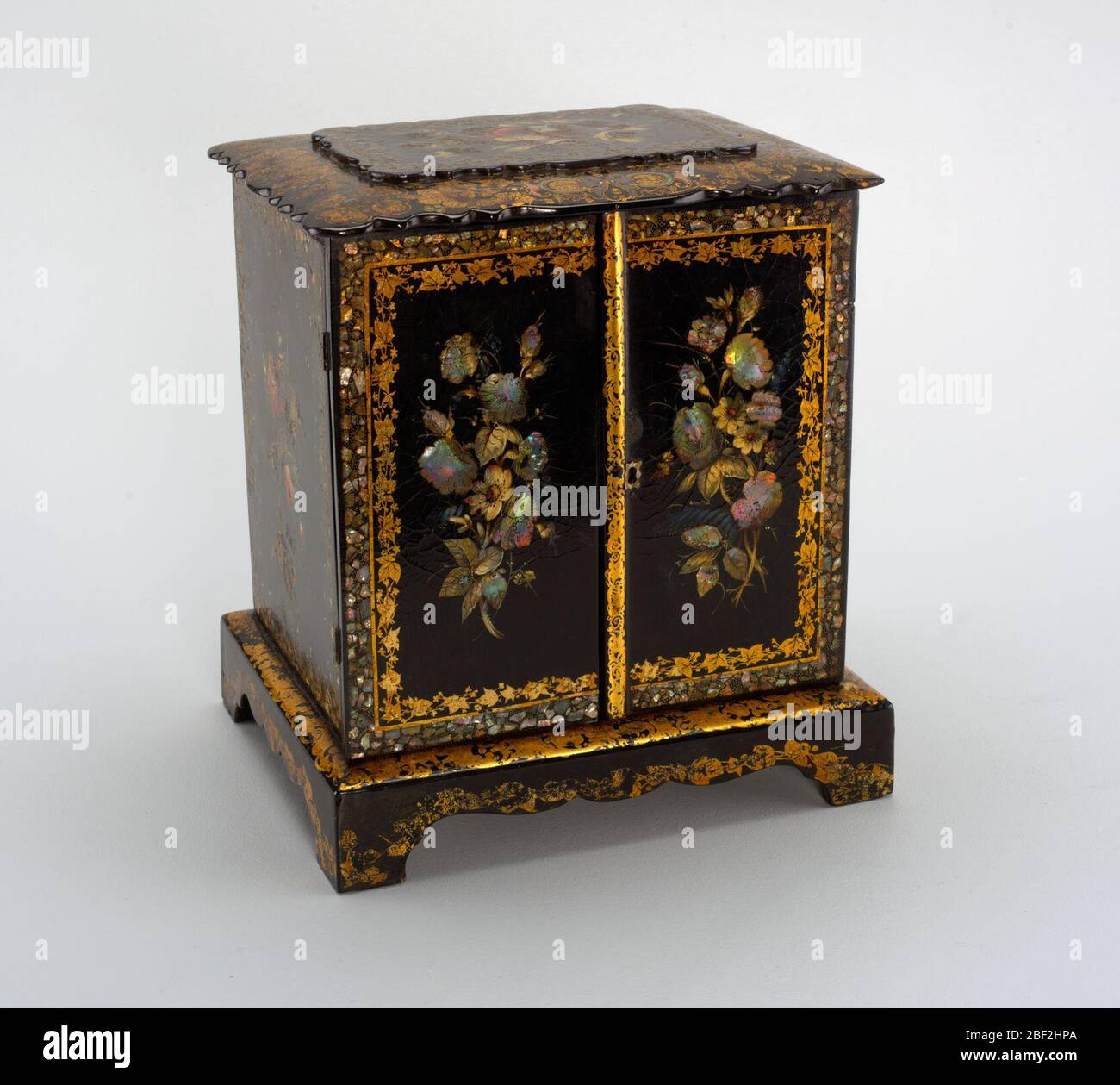 Sewing cabinet. Cabinet with two doors; front painted with floral arrangements on black background, surrounded by two frames, one of gilt foliage and the other of mother-of-pearl. Top with scalloped edge and decorated with colored foliage. Stock Photo