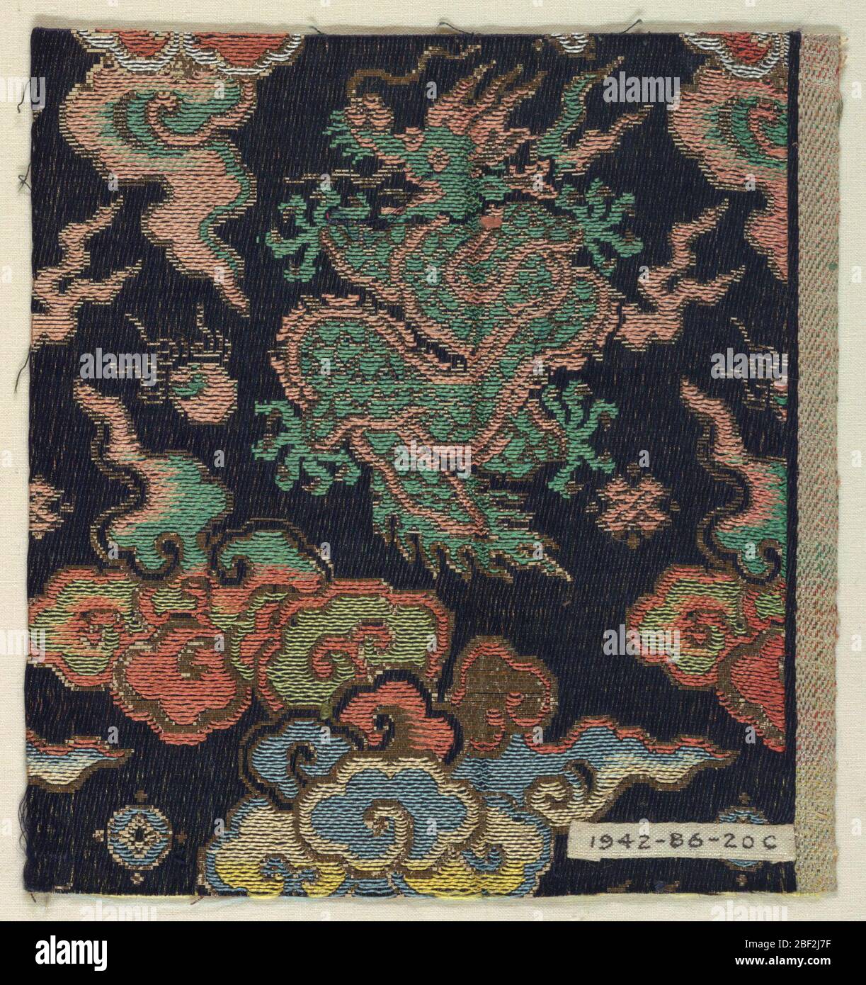 Fragment. Navy blue twill ground with woven supplementary weft pattern of large scale dragons and flying clouds in rust, white, yellow, green, and sky blue silk threads with metallic paper threads. Stock Photo