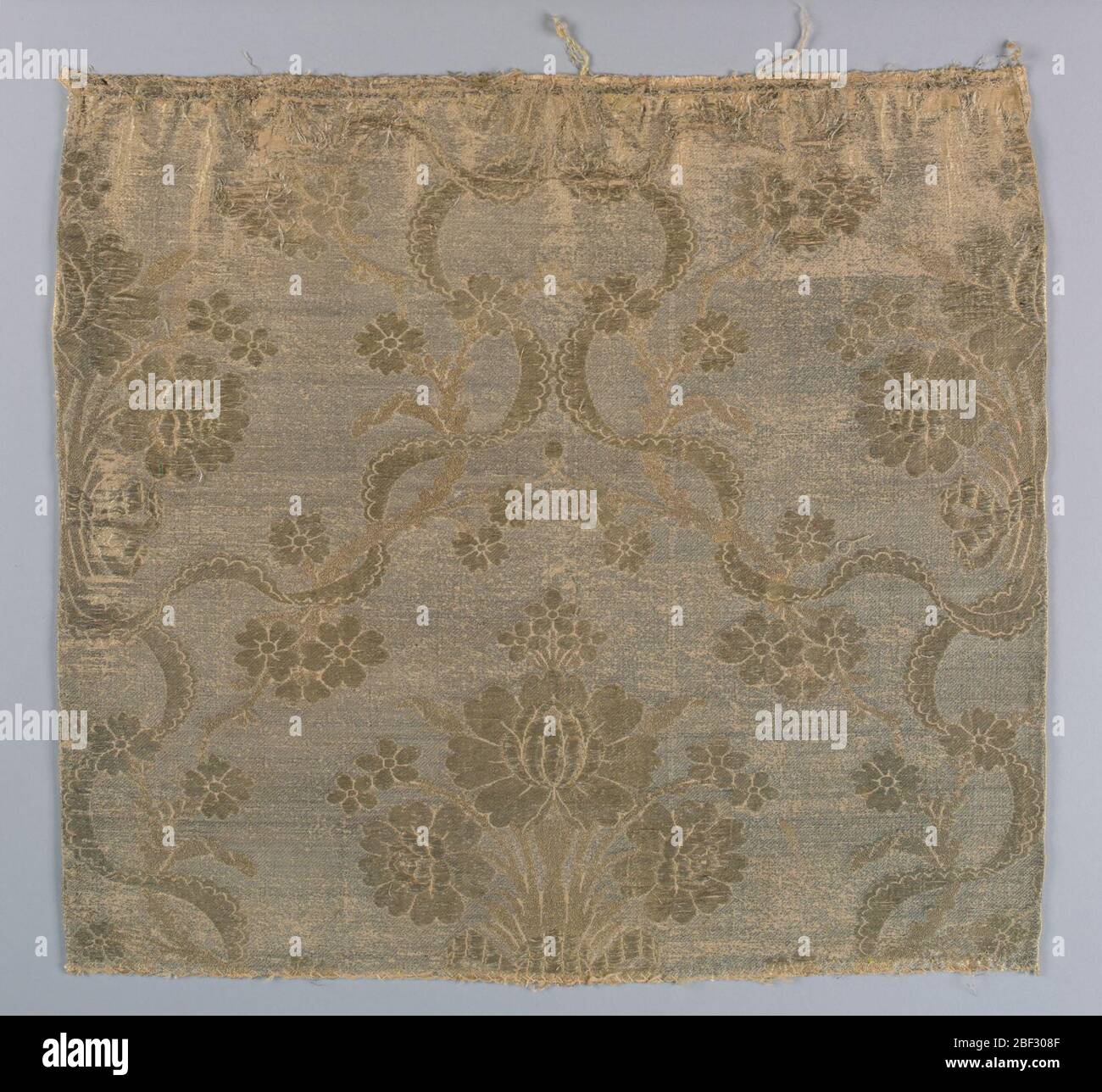 Fragment. Peach and metallic silver fabric in a design of a large-scale central spray of roses flanked by scalloped ribbon serpentines meeting in the center. Large roses in high relief. Metallic brocading in several kinds of gold thread. Stock Photo