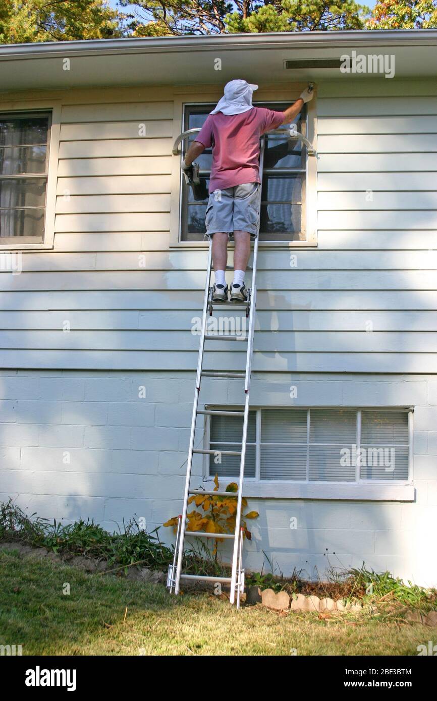 A man precariously balances atop a ladder set on an incline as he paints the exterior of a house on an autumn afternoon Stock Photo
