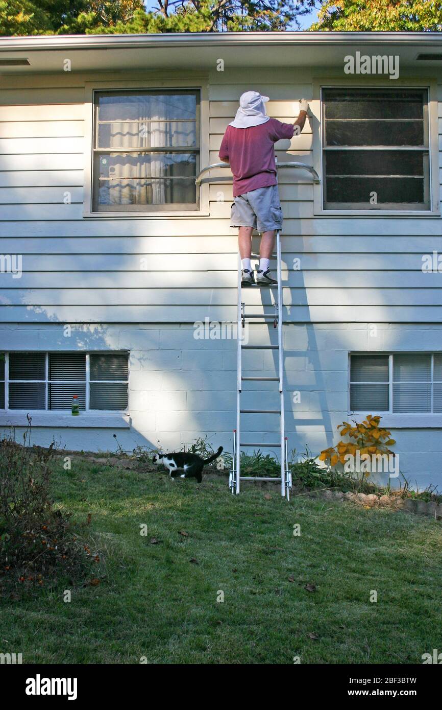 A man precariously balances atop a ladder set on an incline as he paints the exterior of a house on an autumn afternoon Stock Photo
