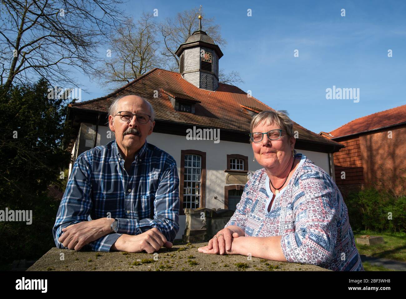 16 April 2020, Lower Saxony, Löwenhagen: The married couple Uwe Kamm (SPD), deputy mayor, and Roswitha Kamm, churchwarden, stand in front of the church of Löwenhagen with its small church tower. Occasionally the bells of the village church in the district of Göttingen do not ring. A raccoon is said to be responsible for this. The animal, which has been staying in the attic of the Protestant church for quite some time, is said to have flipped the lever of the power switch of the bell system. Photo: Swen Pförtner/dpa Stock Photo
