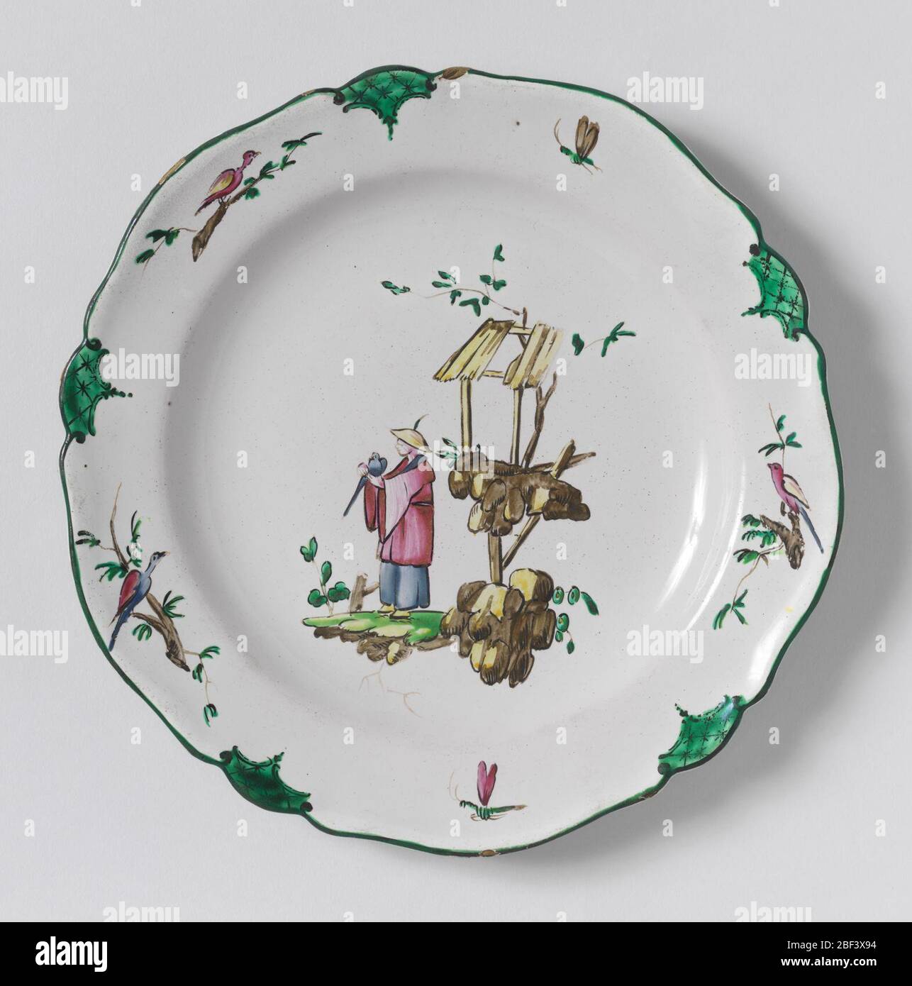 Plate. Circular cavetto, scalloped rim irregularly ten-sided. Chinoiserie decoration showing a manFacing left, holding a birds. At right, rocks and a shelter in brown and yellow. Border has three birds and two flying insects alternating with green scalloped panels. Stock Photo