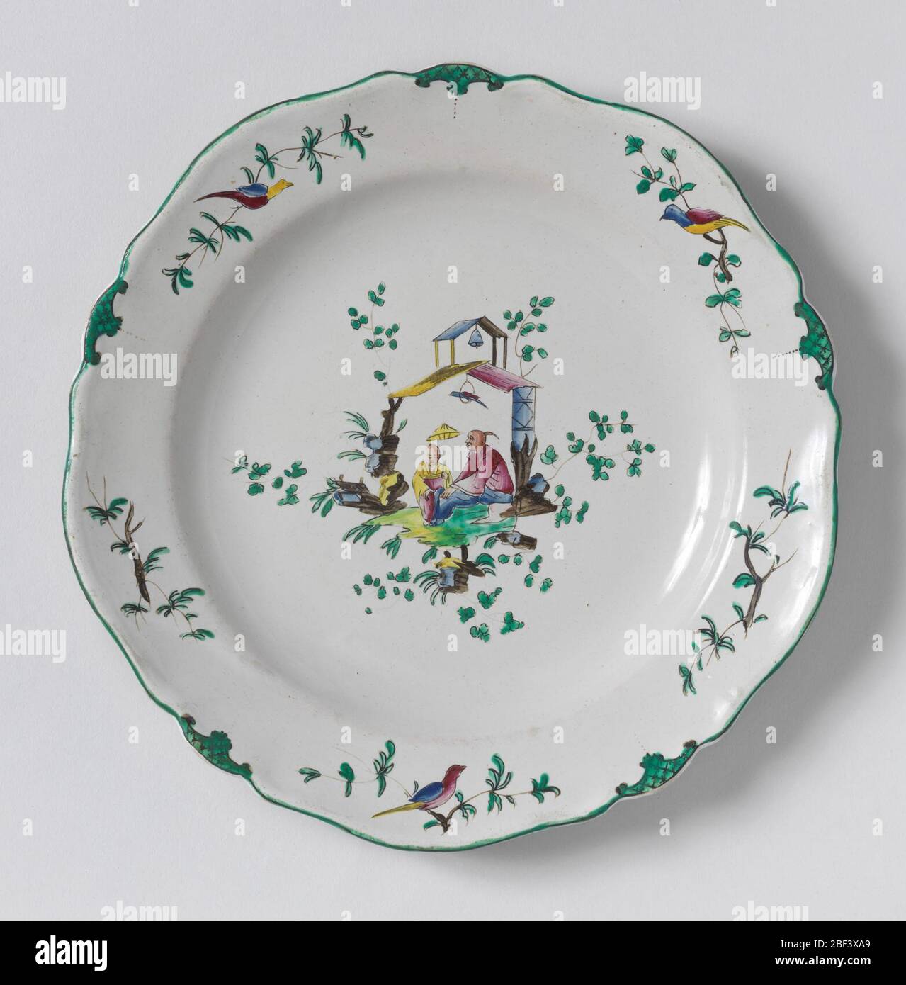 Plate. Circular cavetto, scalloped rim irregularly ten-sided. Chinoiserie decoration: seated man and boy holding parasol under double-roofed shelter supporting a bell and a bird, the whole surrounded by foliage. Border of alternating birds and green latticed panels. Stock Photo
