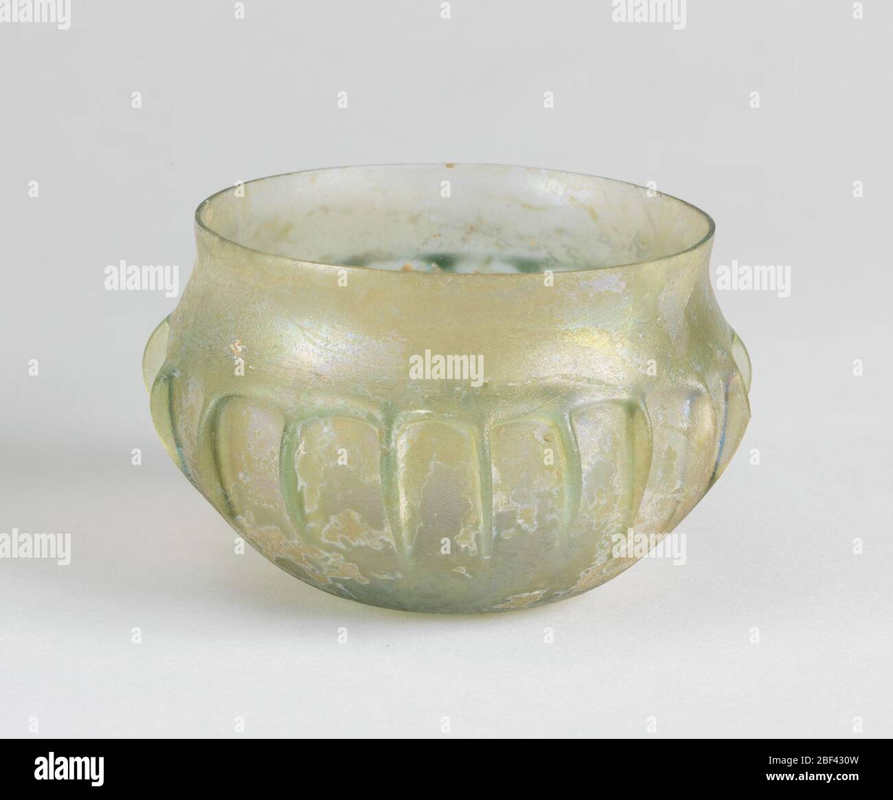 Bowl. Rounded body of greenish glass with iridescence. Vessel has contracting concave neck, continued in eighteen vertical ribs at the center of the vessel's body. Design shows derivation of Lotus-petal design. Stock Photo