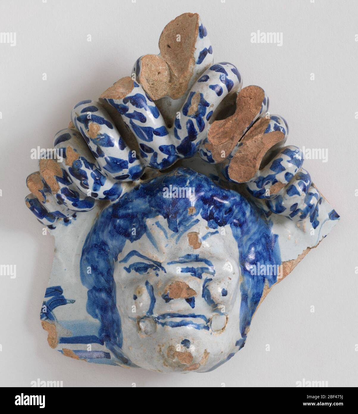 Fragment. Mascaron, crowned with coiled serpents. Pale blue glaze over pink earthenware body, with details in dark cobalt. Stock Photo
