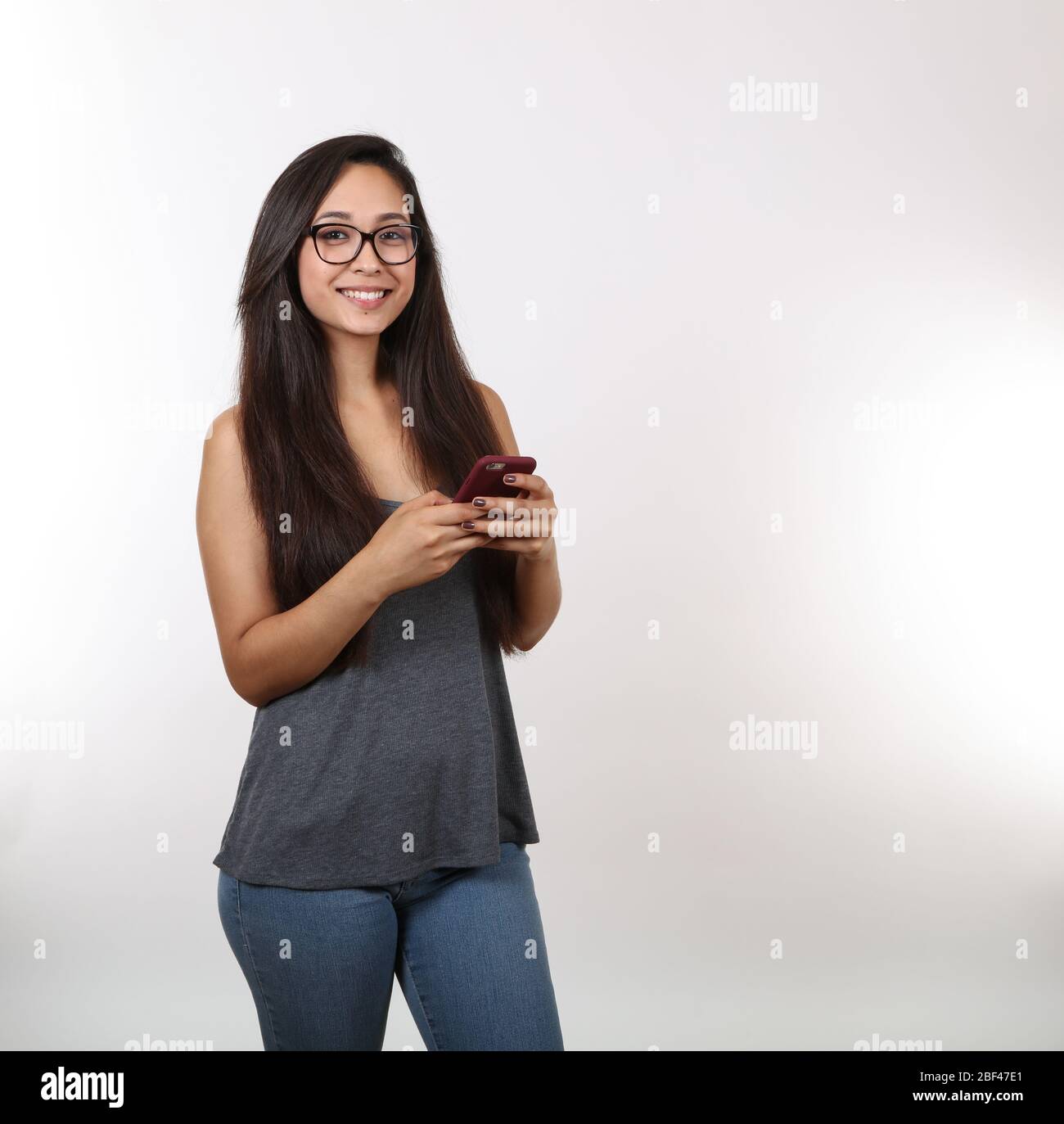 A young lady wearing glasses, blue jeans and a grey tank top holds her phone as she smiles. Stock Photo