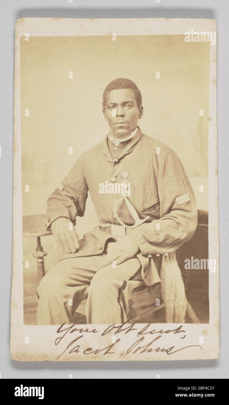 Carte de visite of Sgt Jacob Johns 19th USCT. This carte-de-visite has a photographic print depicting Sergeant Jacob Johns seated, facing the viewer with his gloved hands resting on his lap. His right elbow is resting on a small, circular side table, while it appears he is holding his left arm up without support. Stock Photo