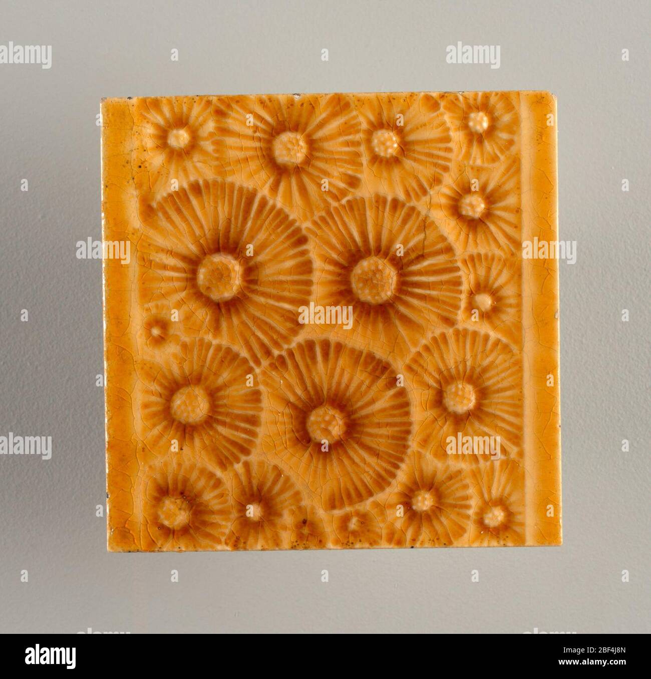 Tile. Square molded tile made of white clay, Tile face is decorated with stylized dandelions (gone to seed) in an all-over pattern set between plain horizontal borders. Tile is painted deep honey-brown crackle glaze. Stock Photo