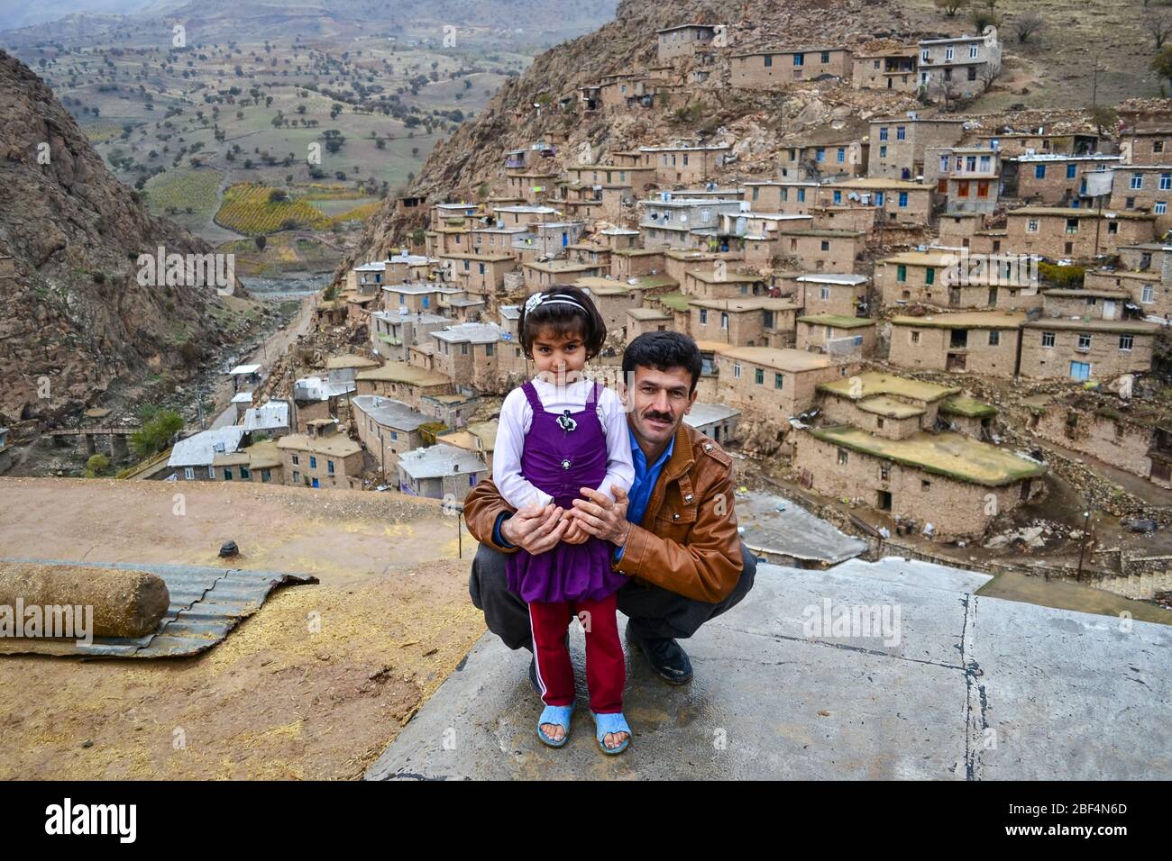 Palangan, Iranian Kurdistan - November 15, 2013: Portrait of cute and little Kurdish girl huged by her father while posing with Palangan stone village in the background Stock Photo