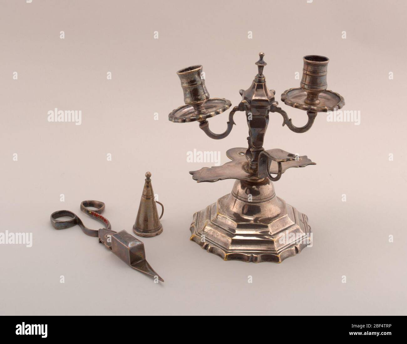 Candlestick with snuffers. Chamber candlestick with stepped base, scalloped at bottom. Two s-curve branches support ringed sockets. Finial at center. Ring handle with thumb rest. Candle snuffer is a small hollow cone with ball finial. Stock Photo