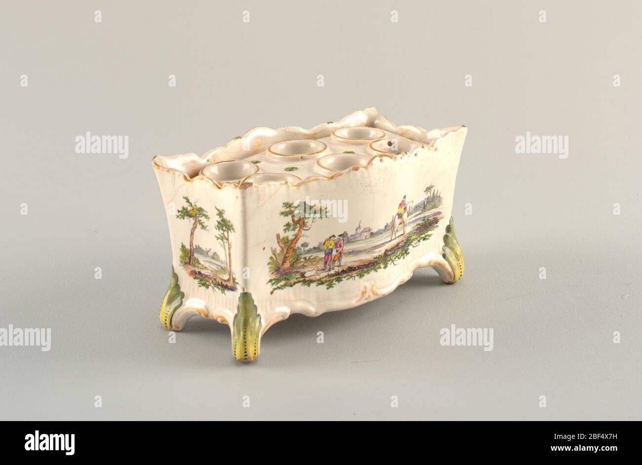Flower pot and cover. Oblong form that flares at top. Scalloped upper and lower edge with traces of gilding. Faint molded ridges. Scrolling acanthus leaf feet painted green. Place for six bulbs. Landscape with figures and dog. Stock Photo