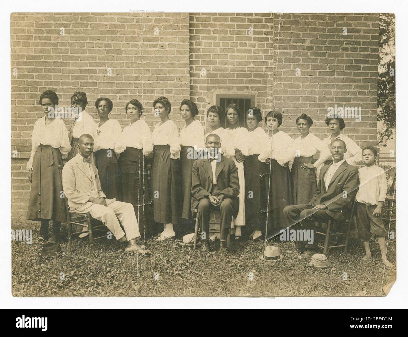 Photograph of Lucille Brown and Elder Brown among others. This black and white photograph depicts twelve women, three men, and one boy. The women all stand a row wearing light-colored shirts and dark skirts, except one woman fifth from the right, who wears a light-colored skirt. The women all face the left and stand at an angle. Stock Photo