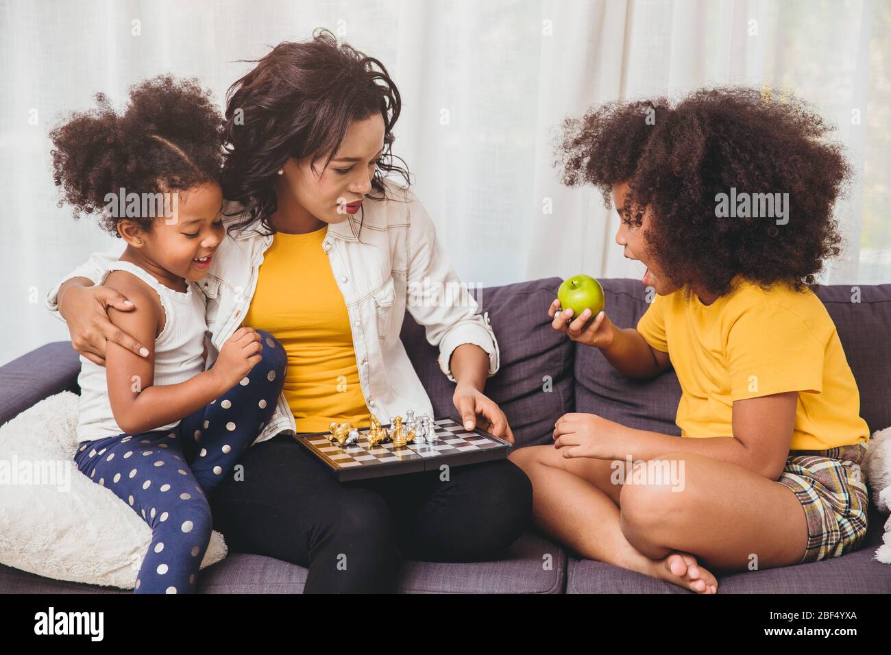 Single mom good care, teaching her childs to be genius and clever little girls learning to play chess boardgame eating fruit for healthy. Stock Photo