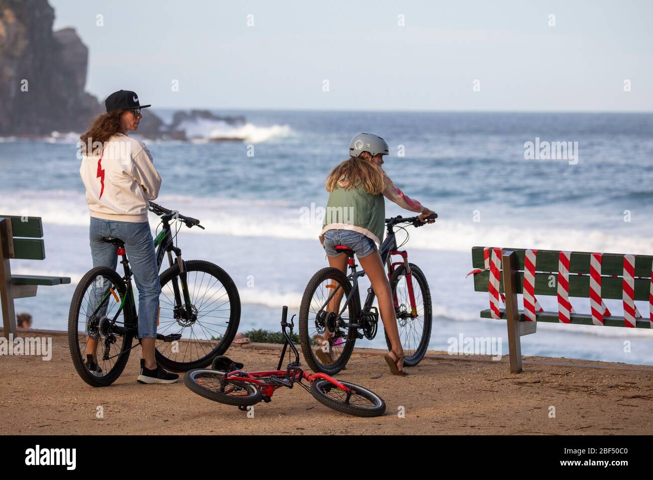 Two teenage girls riding bicycles at the beach in Sydney during covid 19 lockdown with benches taped off to prevent people loitering,Sydney,Australia Stock Photo