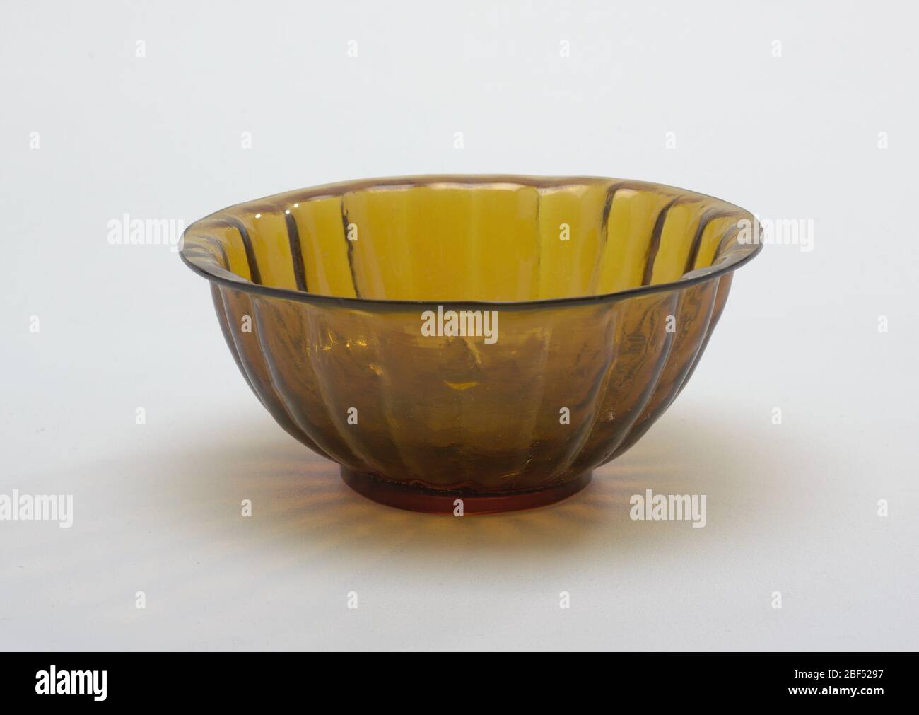 Bowl. Mold-blown bowl of amber colored glass with an inverted rim, a ring foot, low and flat; sloping sides of radiating petal shaped flutes. Interior of bowl is smooth while glass on exterior is rough. Base polished flat, parallel striations on exterior sides. Stock Photo