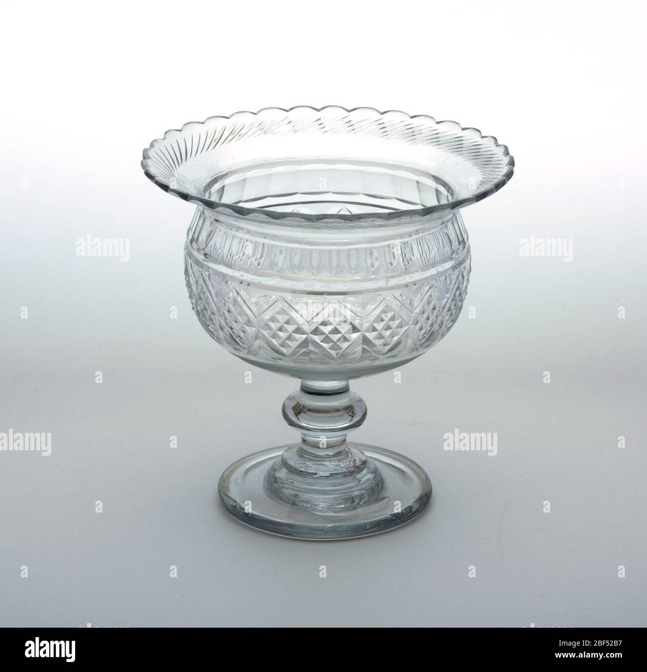 Bowl. Spherical bowl with wide flaring rim. Stem with flattened knop and stepped circular base. Top rim has scalloped edge and row of cut blazes. Stock Photo