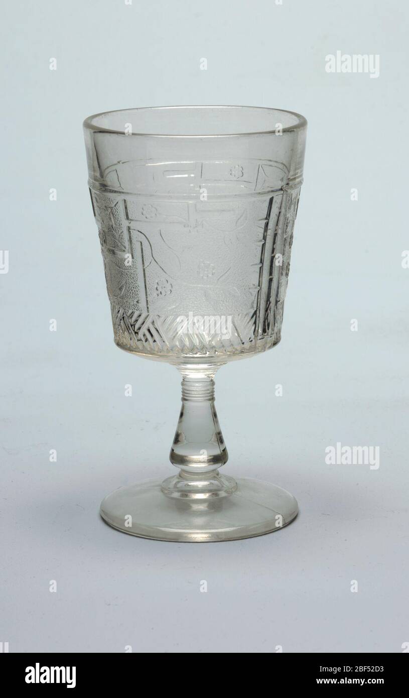 Goblet. Clear glass. Tapering cylindrical cup on flaring conical stem on stepped circular foot. Cup molded with Japanese-inspired decoration of fret work, flowers, and flowing stems and leaves on a pebble-textured ground above a broad band of hatched lines. Stock Photo