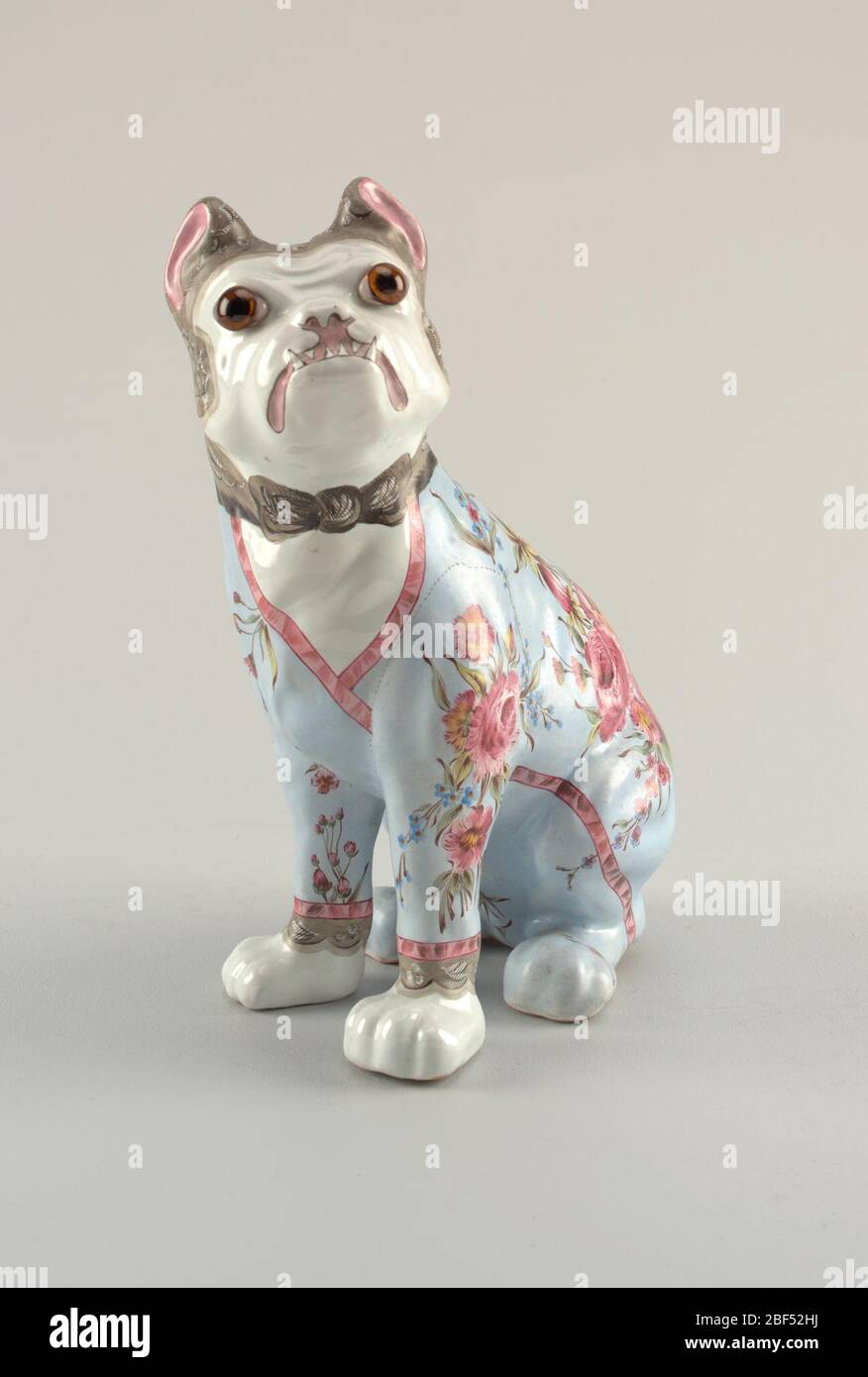 Figure of a dog. Bulldog sitting on haunches, decorated as if wearing light blue dressing gown, banded in red and embellished with predominantly red flowers, a few blue forget-me-nots and green foliage. Stock Photo