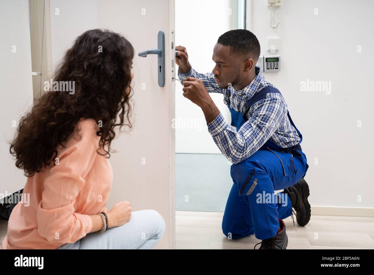 Smiling Woman Looking At Technician Fixing The Door Lock With Screwdriver At Home Stock Photo