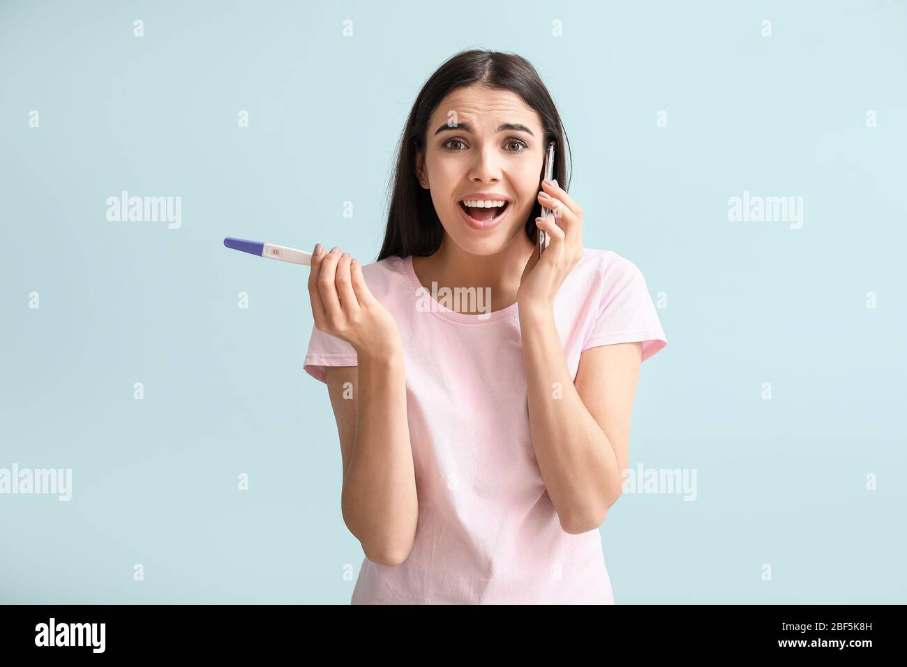 Emotional young woman with pregnancy test talking by mobile phone on