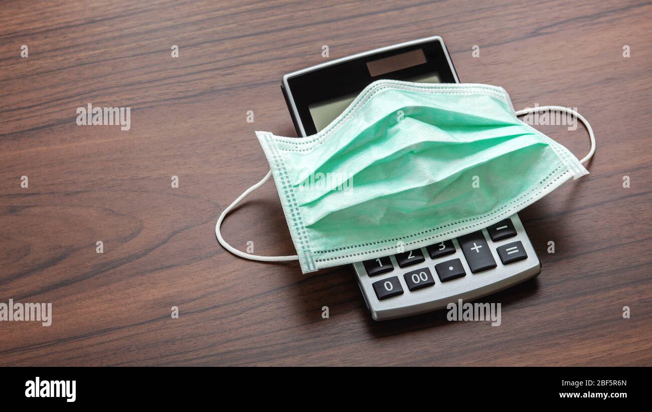 Face mask on calculator on wooden table. Business impact of coronavirus pandemic concept. Stock Photo