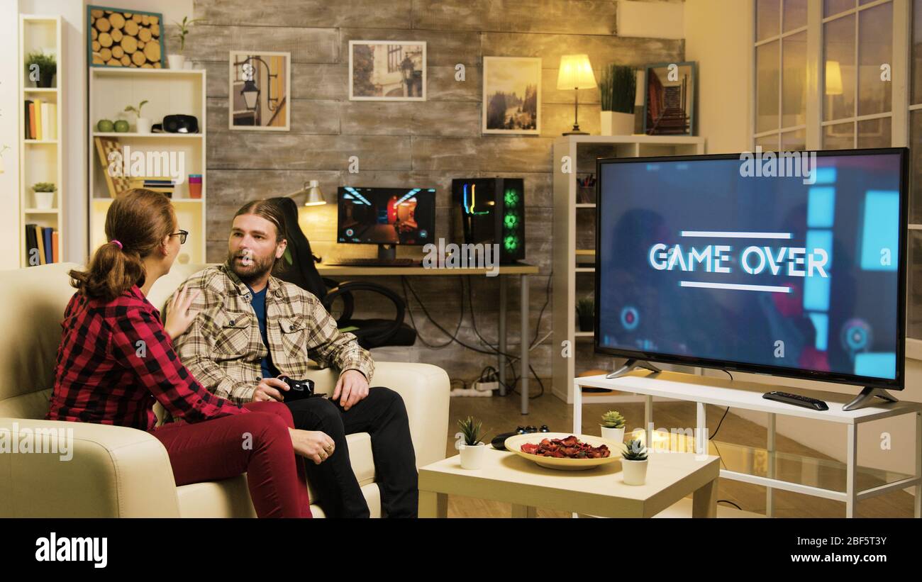 Girlfriend pointing at tv screen while boyfriend is playing video games. Couple sitting on sofa. Stock Photo