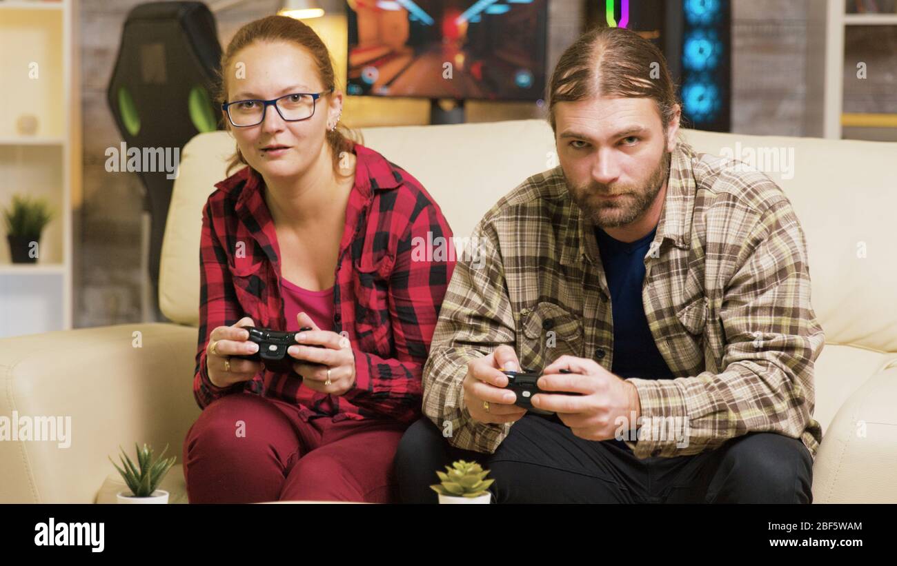 Focused couple playing online video games sitting on couch in living room. Playing games using wireless controllers. Stock Photo