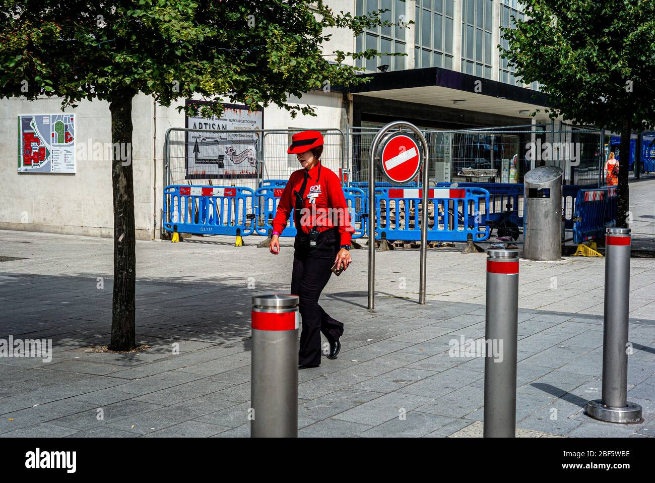 A woman in a red uniform walks down the street past traffic control bollards Stock Photo