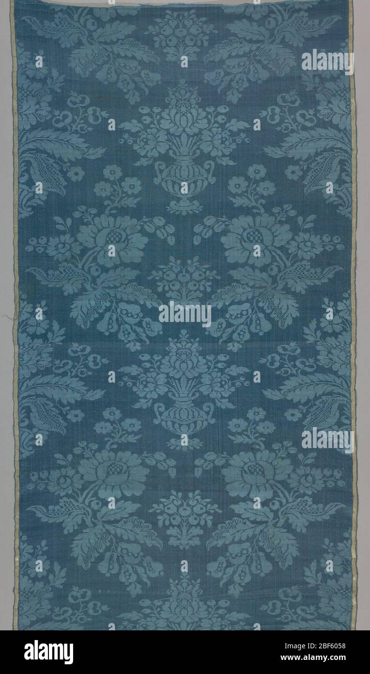 Fragment. Mazarin blue damask with design of lateral rows of curving peony branches with diapered foliage alternating with floral vases. Faded pink and white satin selvages. Stock Photo