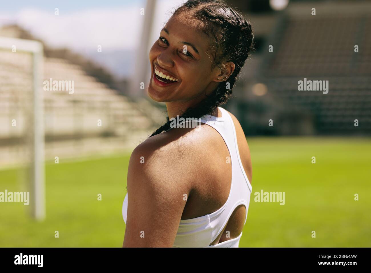 Close-up of a fit female athlete looking over her shoulder and smiling. Woman in sports clothing looking at camera and smiling at sports field. Stock Photo