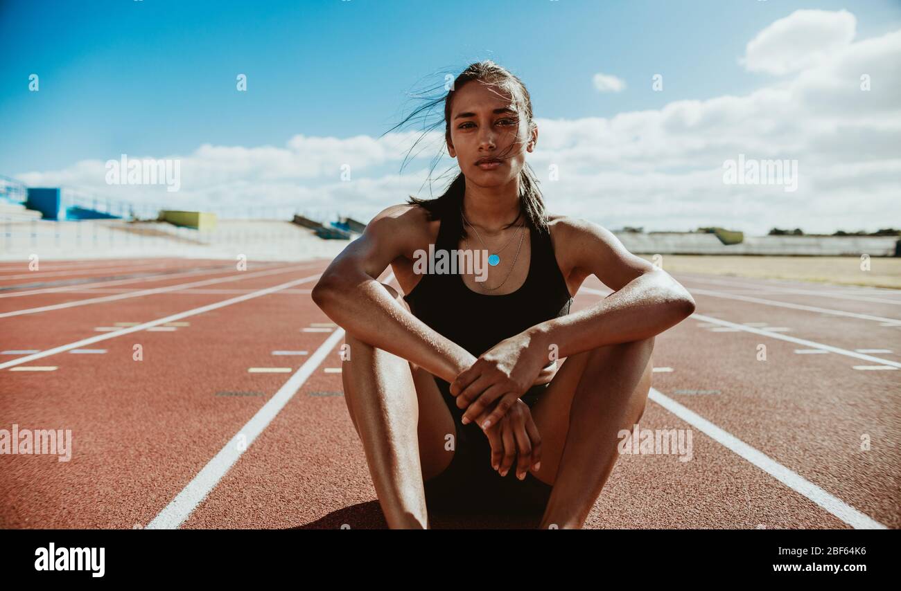 Woman athlete sitting on the track and field stadium. Runner relaxing on the running track after training session. Stock Photo