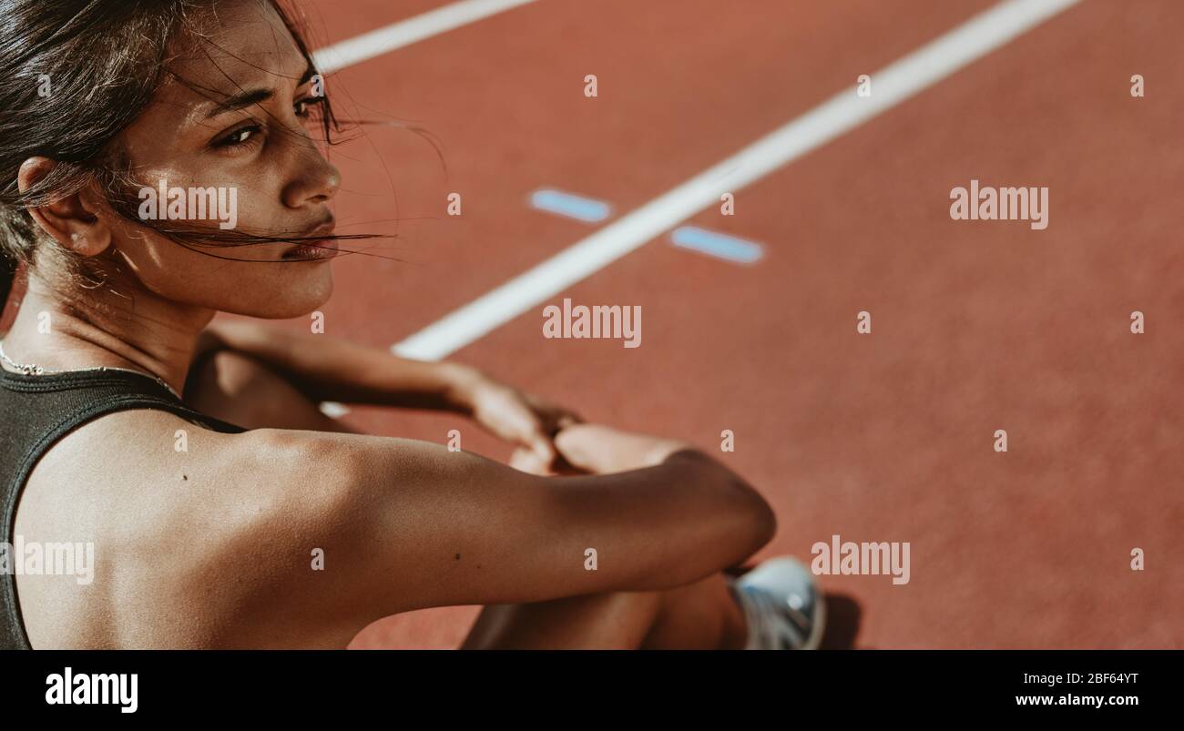 Close-up of a fit woman after run sitting on race track. Determined female athlete sitting on the field and looking away. Stock Photo