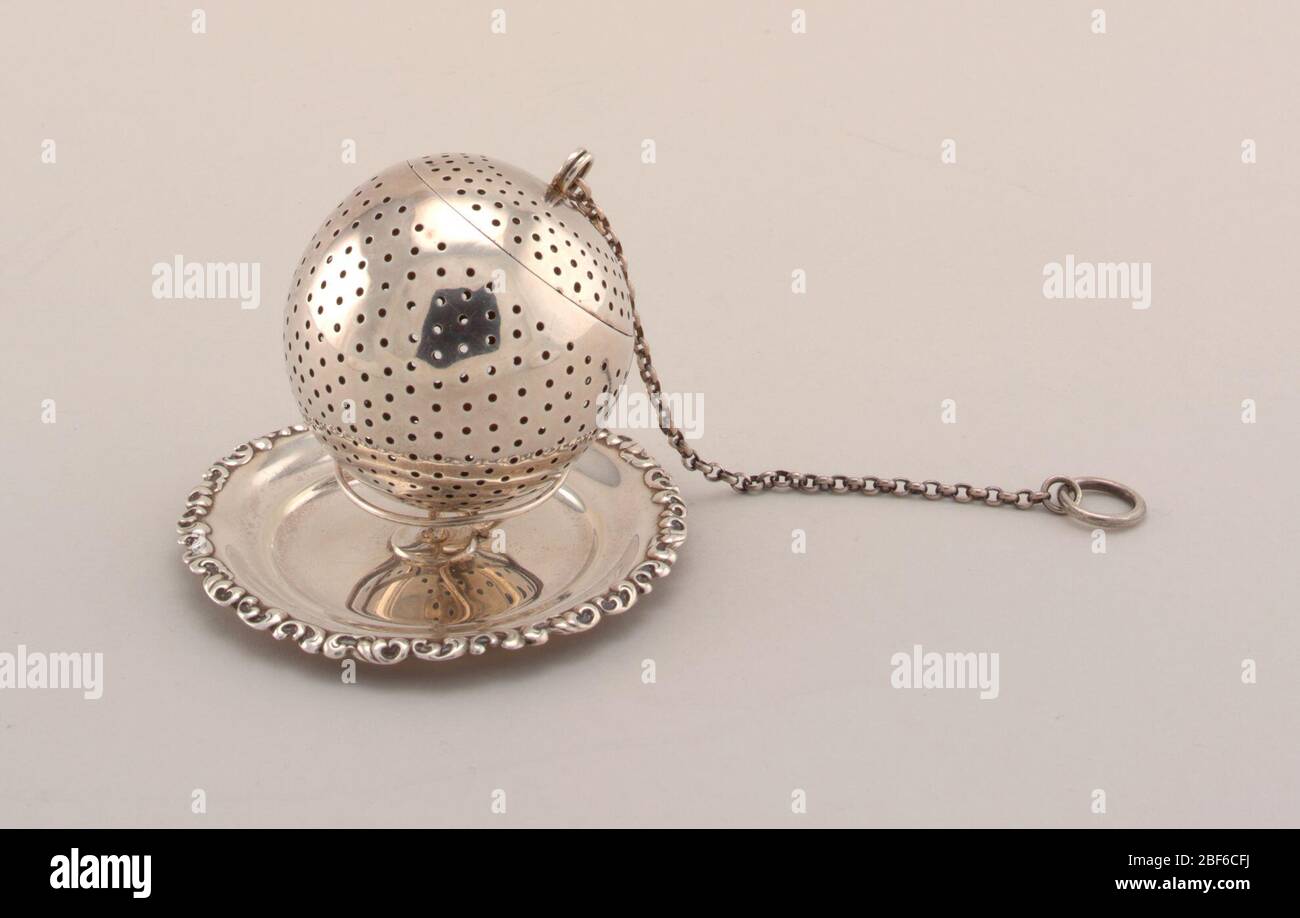 Tea Infuser with Stand. Orb-shaped infuser with chain; stand with wire work support on circular dish with scroll border. Stock Photo