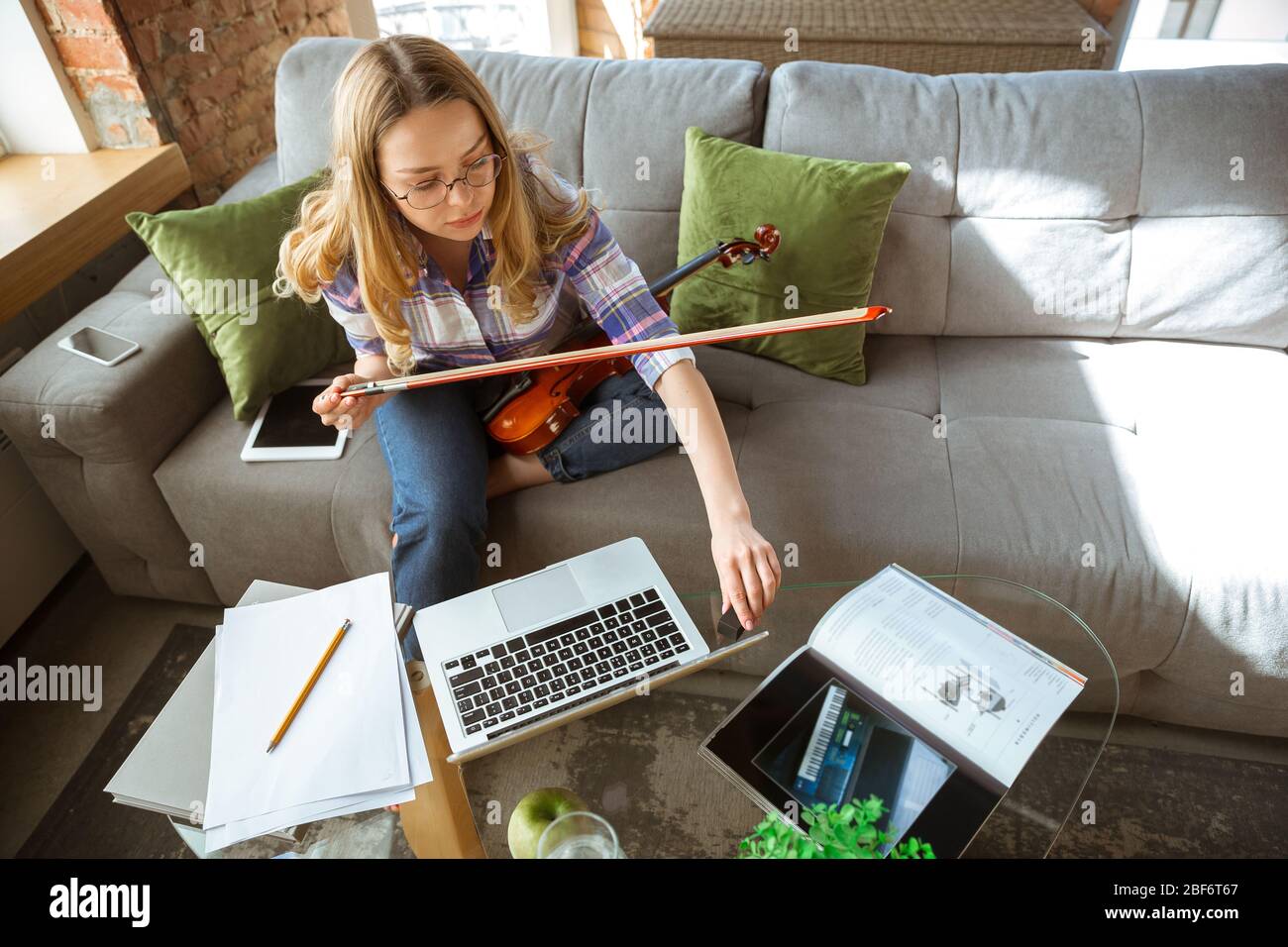 Young woman studying at home during online courses or free information by herself. Becomes musician, violinist while isolated, quarantine against coronavirus spreading. Using laptop, smartphone. Stock Photo