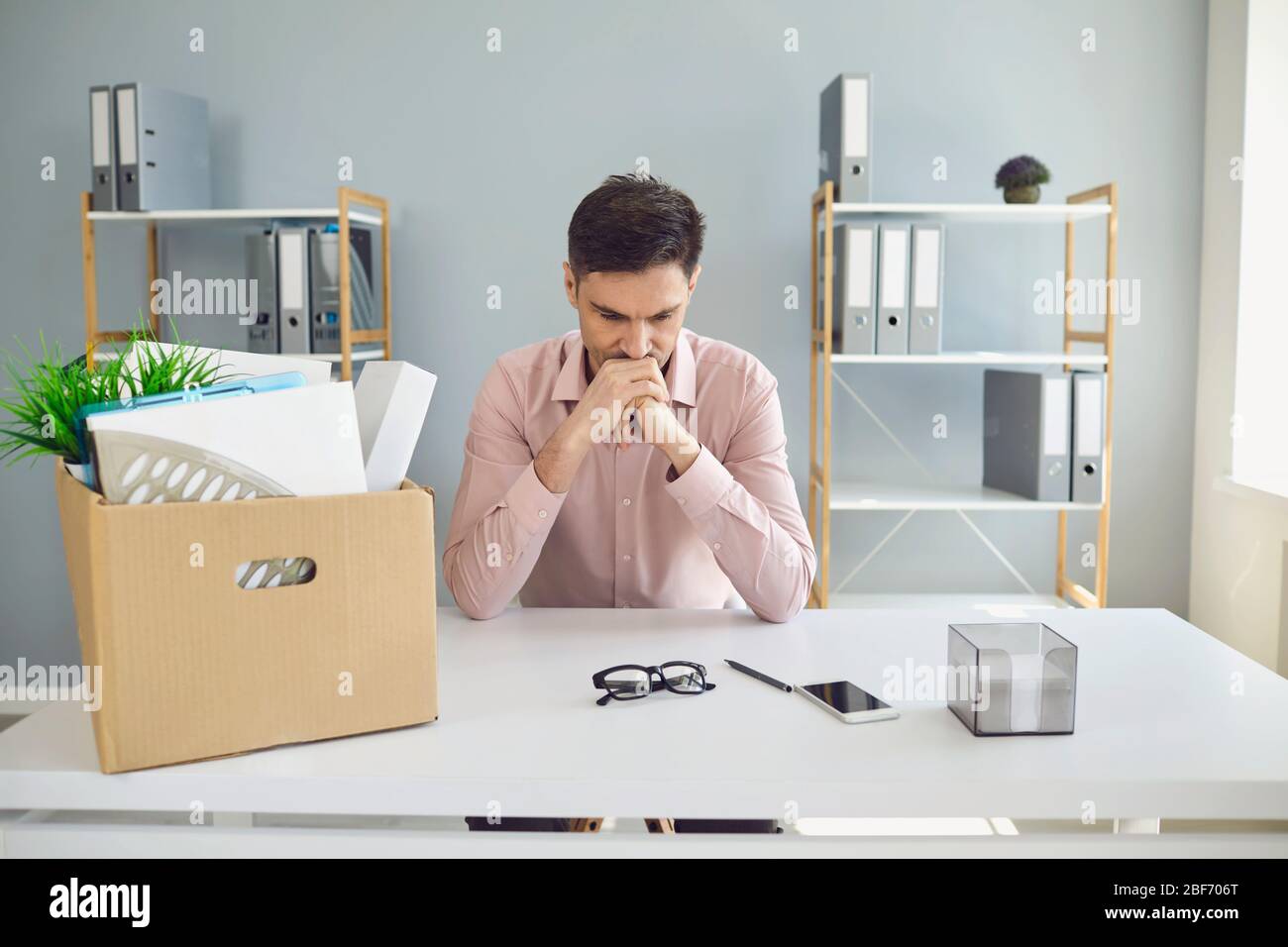 Unemployment. Dismissal. Dismissed businessman is upset with a cardboard box holding his head in his hands while sitting in a company office. Stock Photo