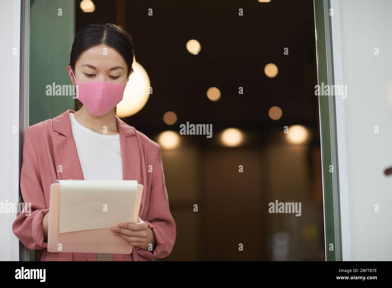 Young Asian woman wearing protective mask standing and making notes in documents she working during epidemic Stock Photo