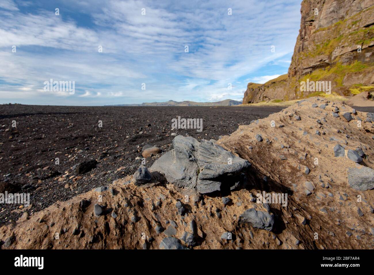 A close-up view of breccia, with chunks of volcanic rock in a light-brown matrix; volcanic sand, cliffs, and a partly cloudy sky are in the background Stock Photo