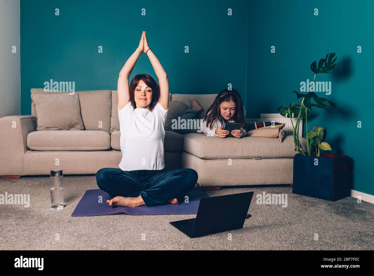 Woman is doing online yoga with laptop during self isolation at her living room, no equipment workout, meditation tips for beginners. Her daughter is reading. Family time with kids, stay home. Stock Photo