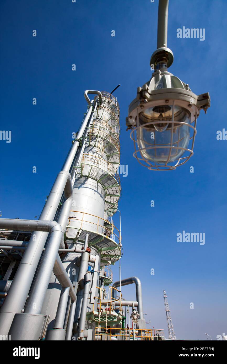 Petrochemical plant. Oil distillation tower on blue sky,  pipes and pipelines. Explosion-proof lamp on foreground. Focus on background. Stock Photo