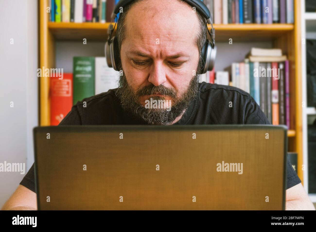 Home office work concept. Adult bearded man concentrated, working online from home on computer laptop, book shelves behind him Stock Photo