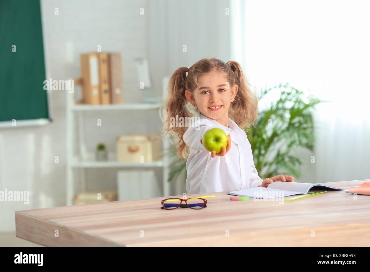Cute little schoolgirl with apple at desk in classroom Stock Photo - Alamy