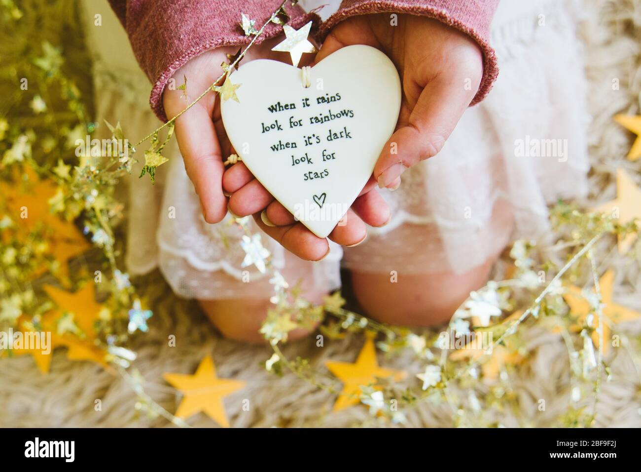 A girl or woman holding a heart with the quote when it rains look for rainbows, surrounded by gold stars. Coronavirus lockdown positive uplifting Stock Photo