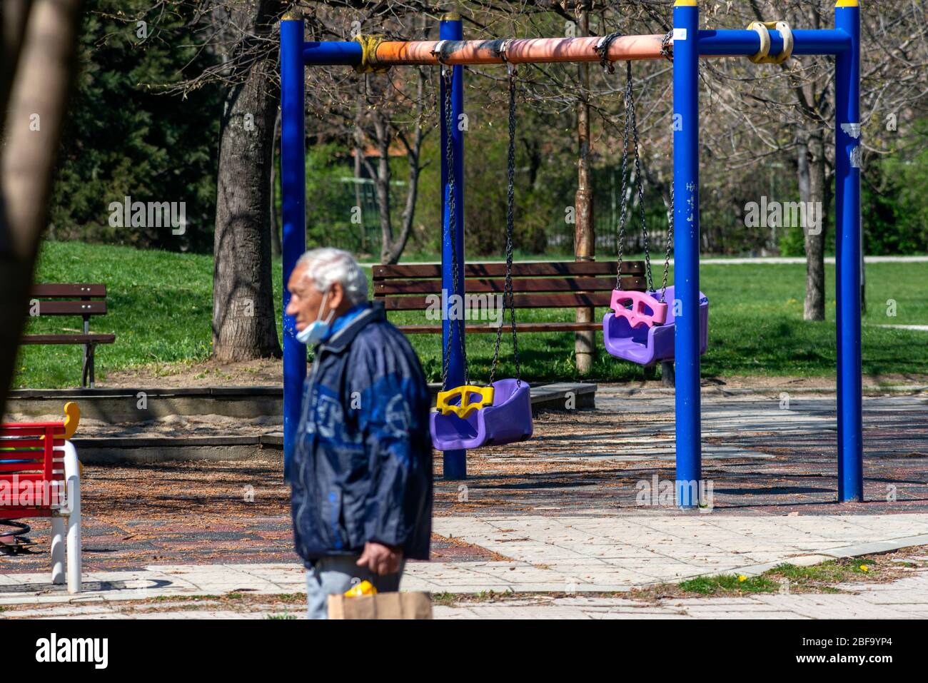 Sofia, Bulgaria. 16 April 2020. Senior man wearing protective face mask walking past empty deserted blue swings at colourful playground or play area on beautiful sunny day. The public park playgrounds have been closed during the lockdown because of the Coronavirus Pandemic of Covid-19 spread. Stock Photo