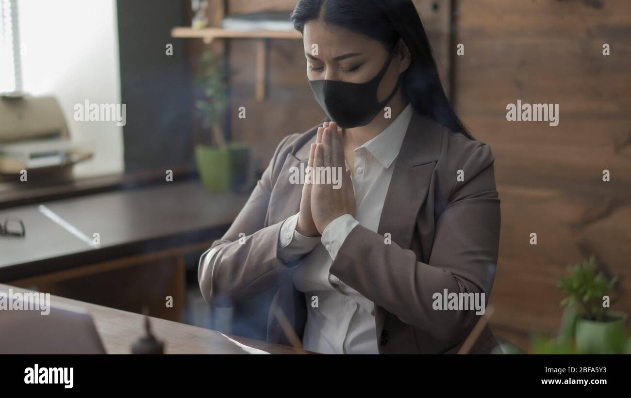Businesswoman In Protective Mask Prays Alone In Office Stock Photo