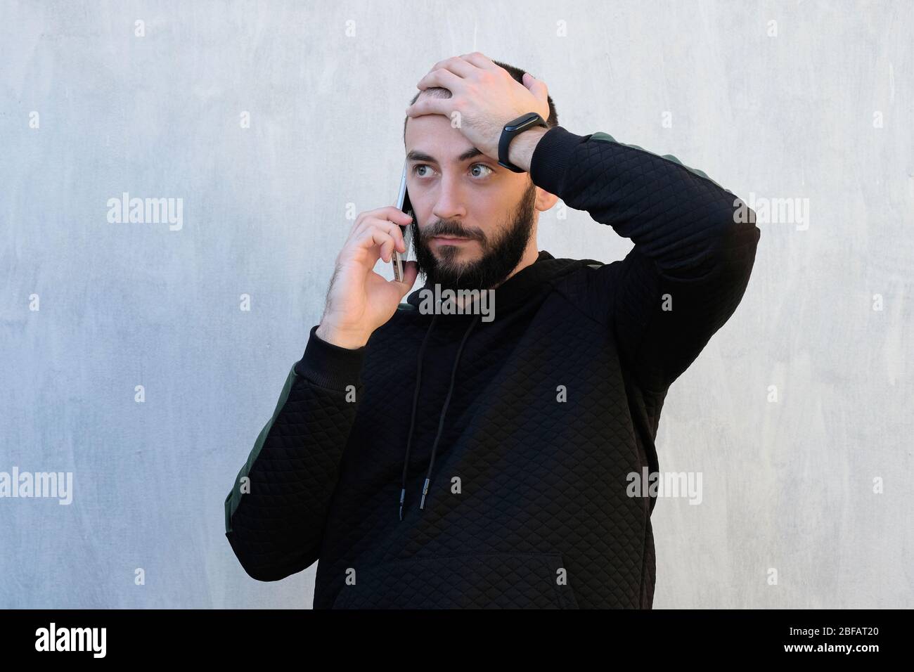 The guy is talking on the phone against a gray wall. The guy holds the phone near his ear and holds his head with other hand. Reporting bad news over Stock Photo