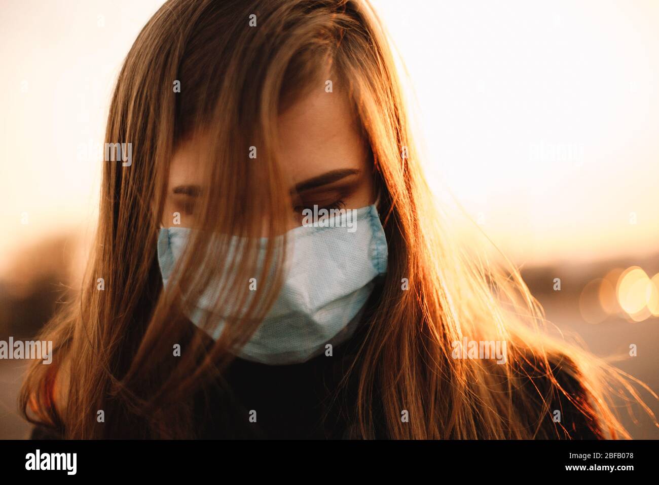 Close up portrait of sad depressed young woman wearing protective face medical mask while standing in city during sunset Stock Photo